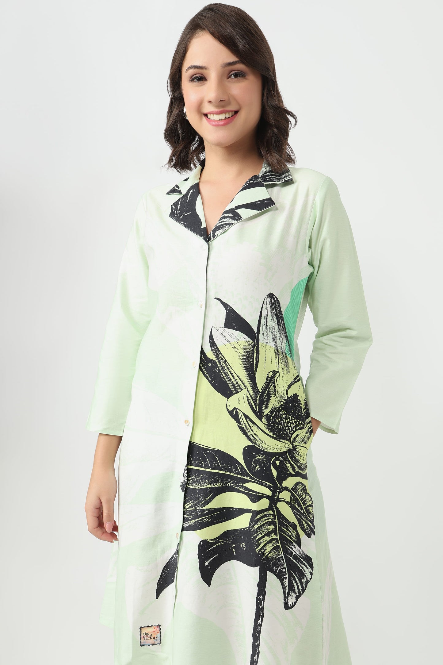 A woman wearing a long-sleeve, collared tunic dress with a light mint green base and a large black botanical leaf print on the front, set against a plain light grey studio background.