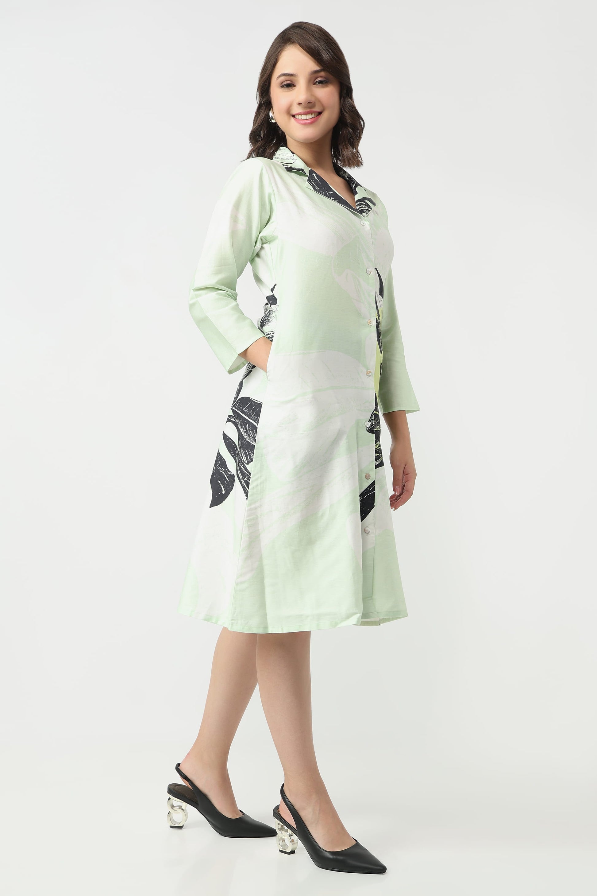 A woman wearing a light green and white tropical print midi dress with black and white leaf patterns, paired with black slingback heels, against a plain white studio background.