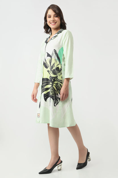 A woman Wearing a light mint green knee-length tunic dress with a large black and lime green tropical leaf print on the front, styled with black pointed-toe heels against a plain light grey studio background.