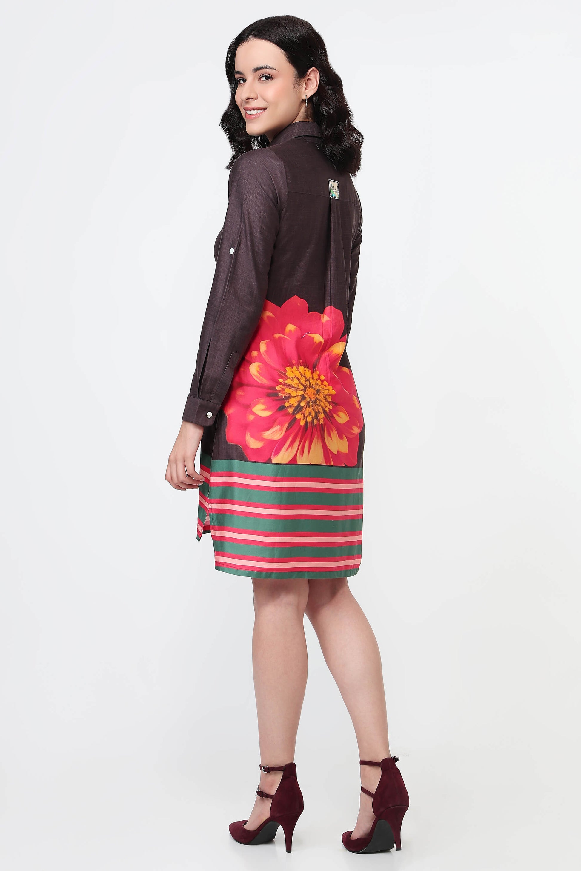 A woman wearing a dark brown long-sleeve shirt dress with a large red and yellow flower print on the back and green and red horizontal stripes on the bottom, paired with black high heels against a white studio background.