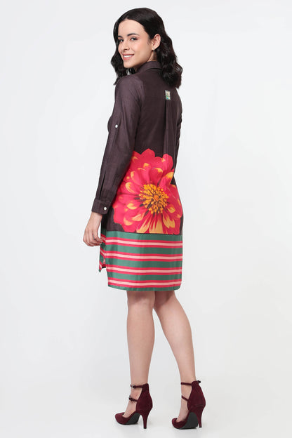 A woman wearing a dark brown long-sleeve shirt dress with a large red and yellow flower print on the back and green and red horizontal stripes on the bottom, paired with black high heels against a white studio background.
