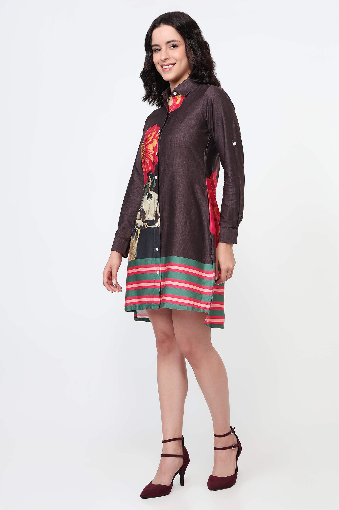 A woman wearing a dark brown long-sleeve shirt dress with a large red and yellow flower print on the back and green and red horizontal stripes on the bottom, paired with black high heels against a white studio background.