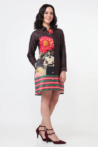 A woman wearing a dark brown long-sleeve shirt dress with a large red and yellow flower print on the back and green and red horizontal stripes on the bottom, paired with black high heels against a white studio background.