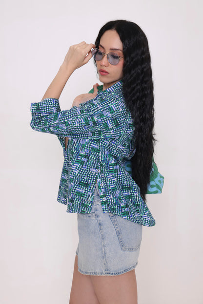 A woman modeling a green and blue geometric patterned shirt paired with a light denim mini skirt, against a plain off-white background.
