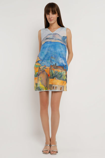 A woman wearing a sleeveless V-neck mini dress with a colourful landscape print in shades of blue, yellow, green, and orange, paired with silver heeled sandals against a plain white background.