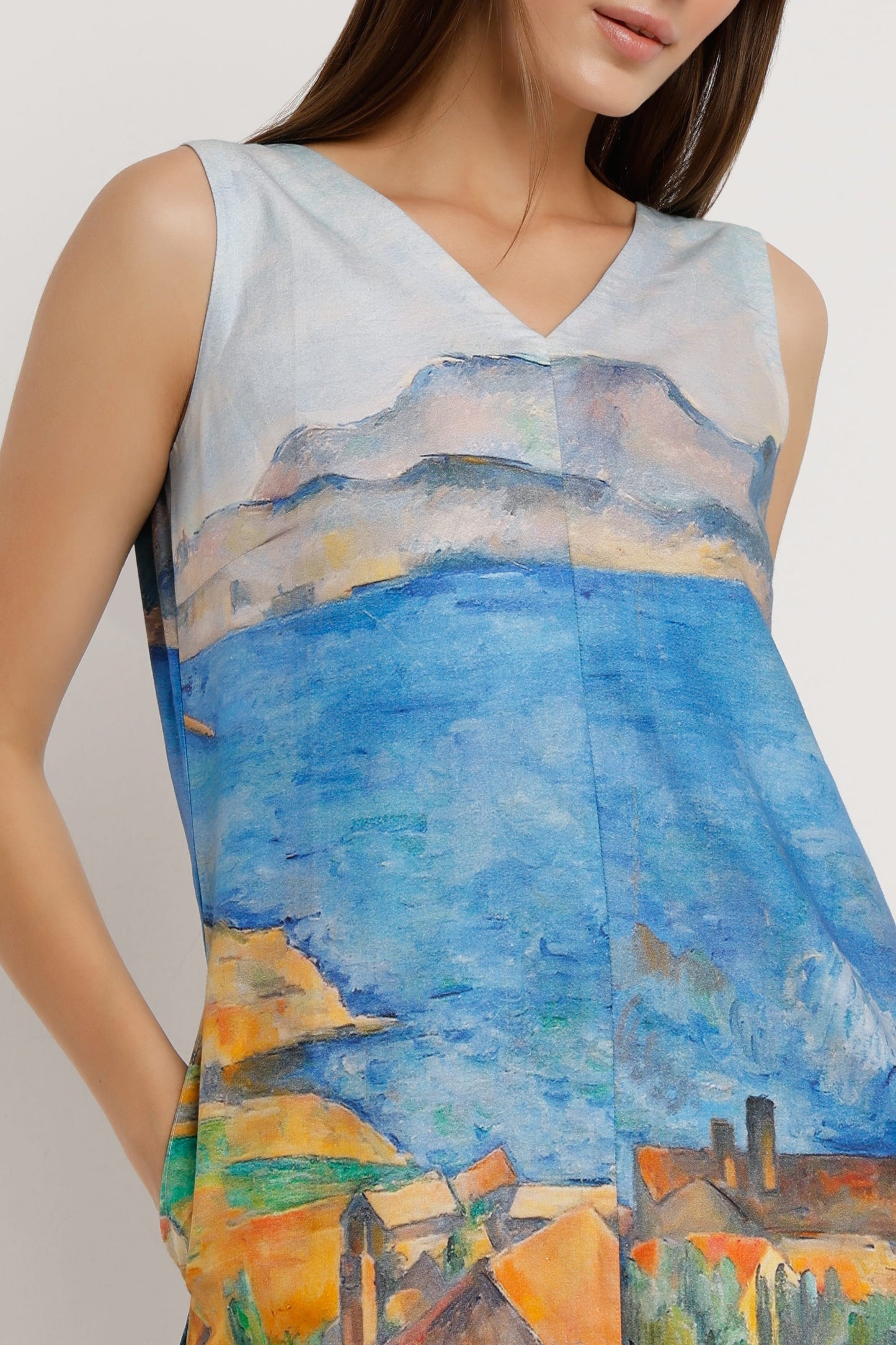 A woman wearing a sleeveless V-neck mini dress with a colourful landscape print in shades of blue, yellow, green, and orange, paired with silver heeled sandals against a plain white background.