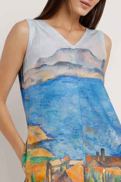 A woman wearing a sleeveless V-neck mini dress with a colourful landscape print in shades of blue, yellow, green, and orange, paired with silver heeled sandals against a plain white background.