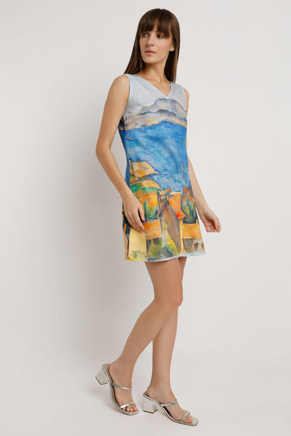 A woman wearing a sleeveless V-neck mini dress with a colourful landscape print in shades of blue, yellow, green, and orange, paired with silver heeled sandals against a plain white background.