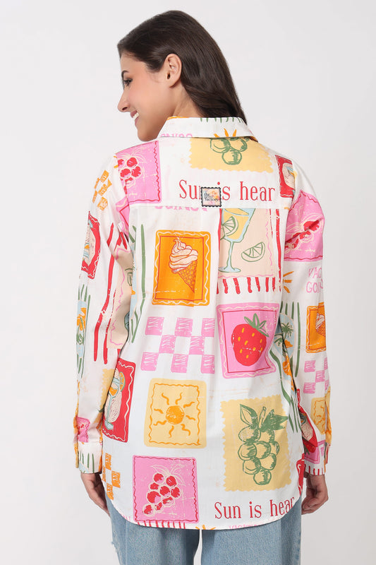 A woman shown from the back wearing a long-sleeve button-up shirt with a colorful fruit and dessert patchwork print on a cream background, paired with light blue jeans against a plain white studio background.