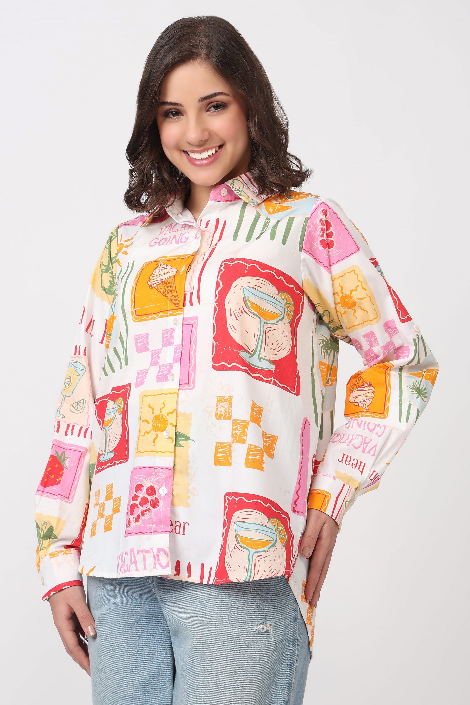 A woman modeling a long-sleeve button-up shirt with a colorful retro patchwork print featuring illustrations of teacups, flowers, and text, paired with light blue jeans against a plain light gray background.