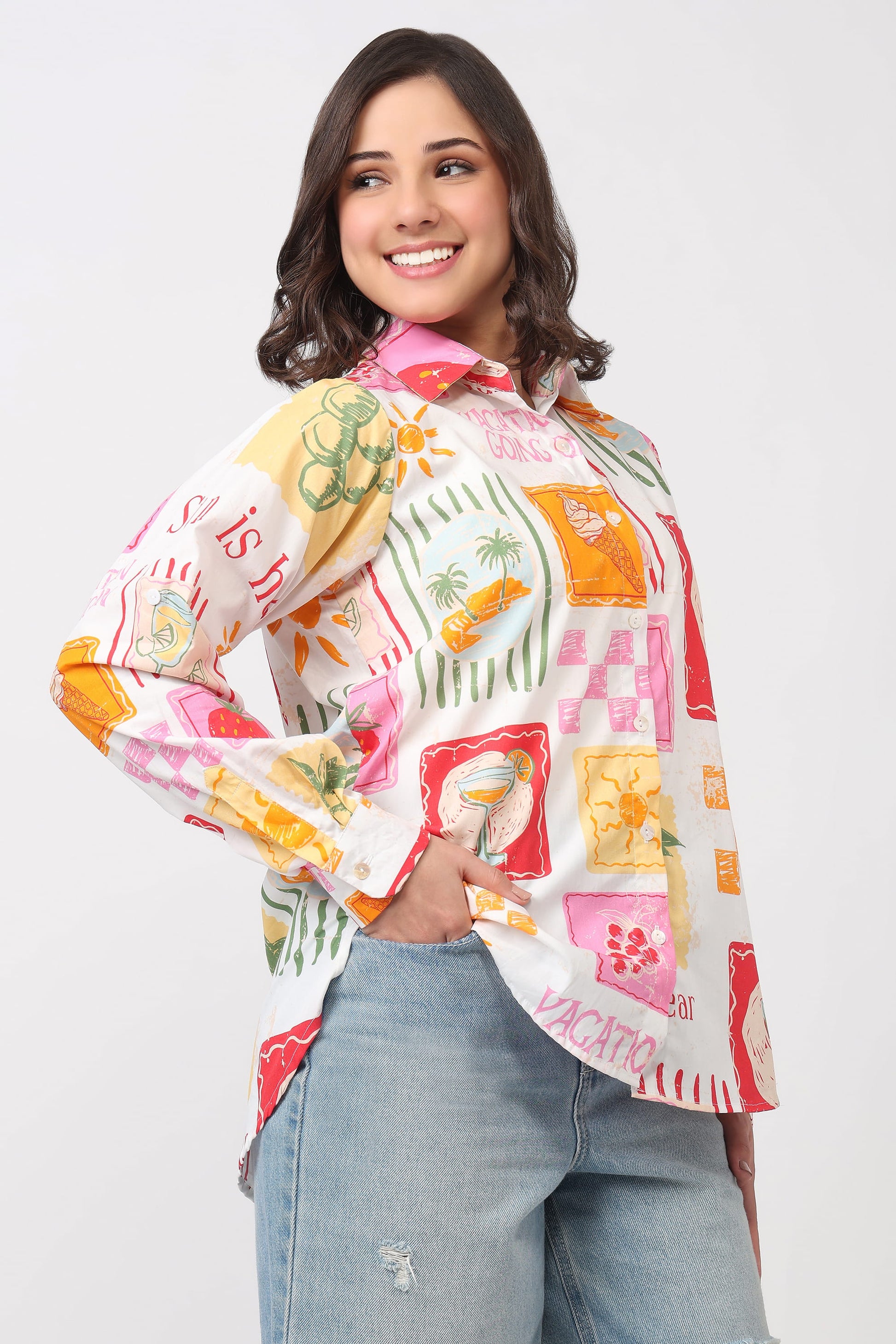A woman wearing a long-sleeve button-up shirt with a colorful tropical patchwork print, paired with light-wash blue jeans, against a plain light gray studio background.