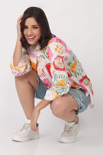 A woman crouching in a studio setting, wearing a white button-up shirt with a colorful tropical and abstract print, denim shorts, and white sneakers.