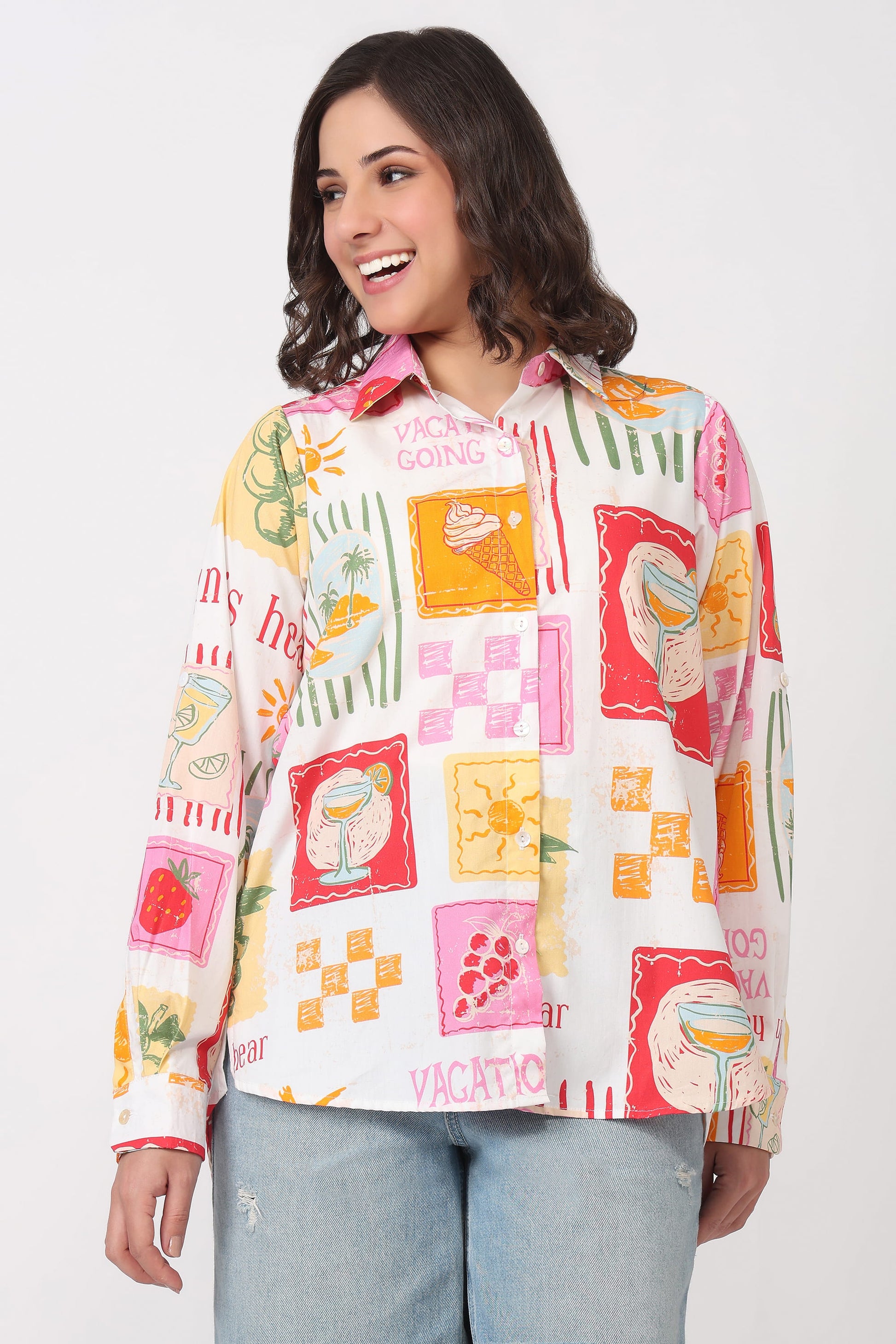 A woman wearing a long-sleeve button-up shirt with a colorful retro travel-themed patchwork print, paired with light-wash jeans, against a plain light gray studio background.
