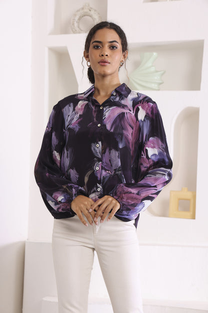 Woman wearing a purple floral blouse and white pants in a minimalistic room.