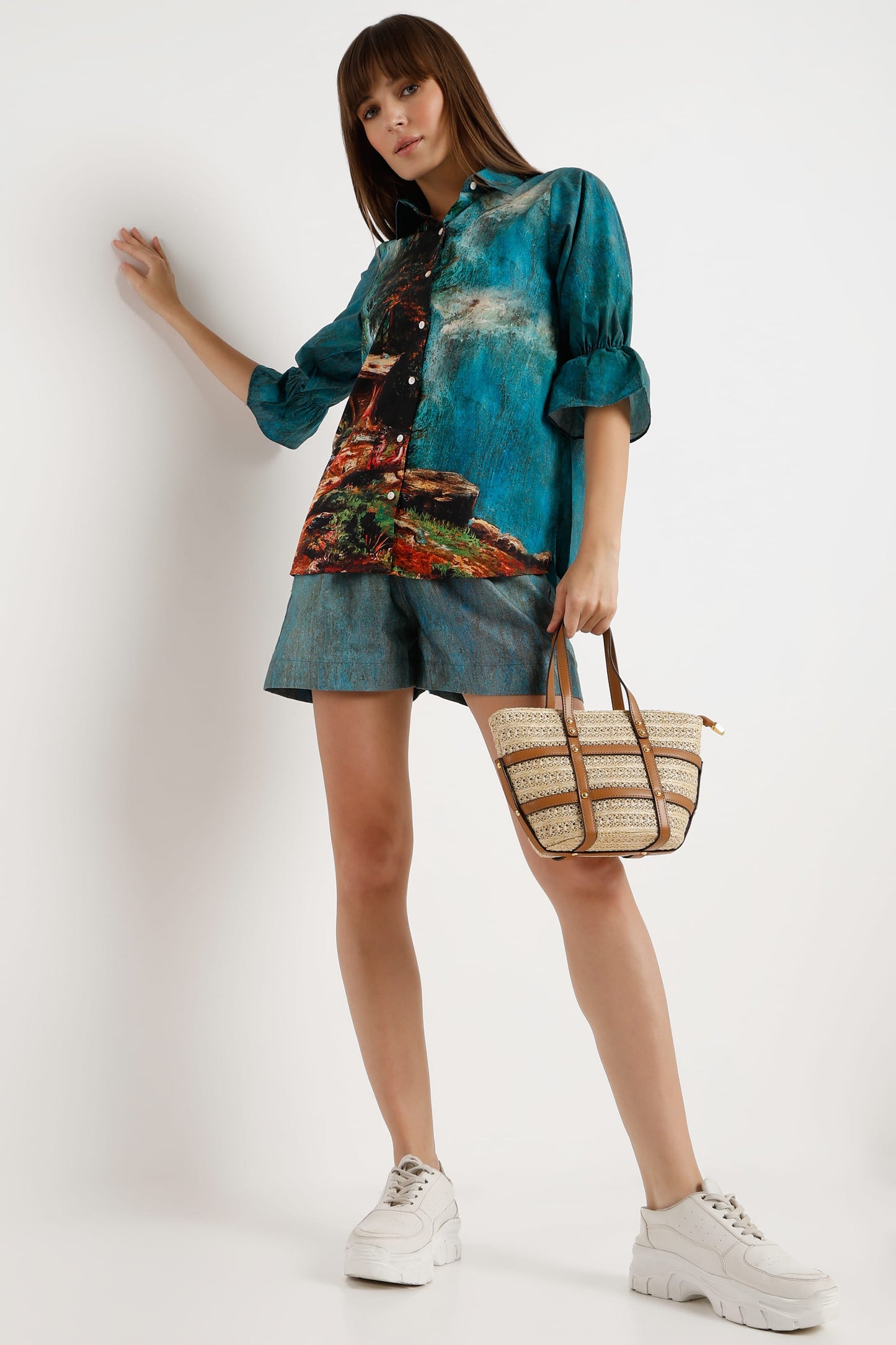 A woman models a matching teal and blue denim-like two-piece set with a colorful abstract print on the shirt, holding a woven straw tote bag and wearing white platform sneakers against a plain white studio background.