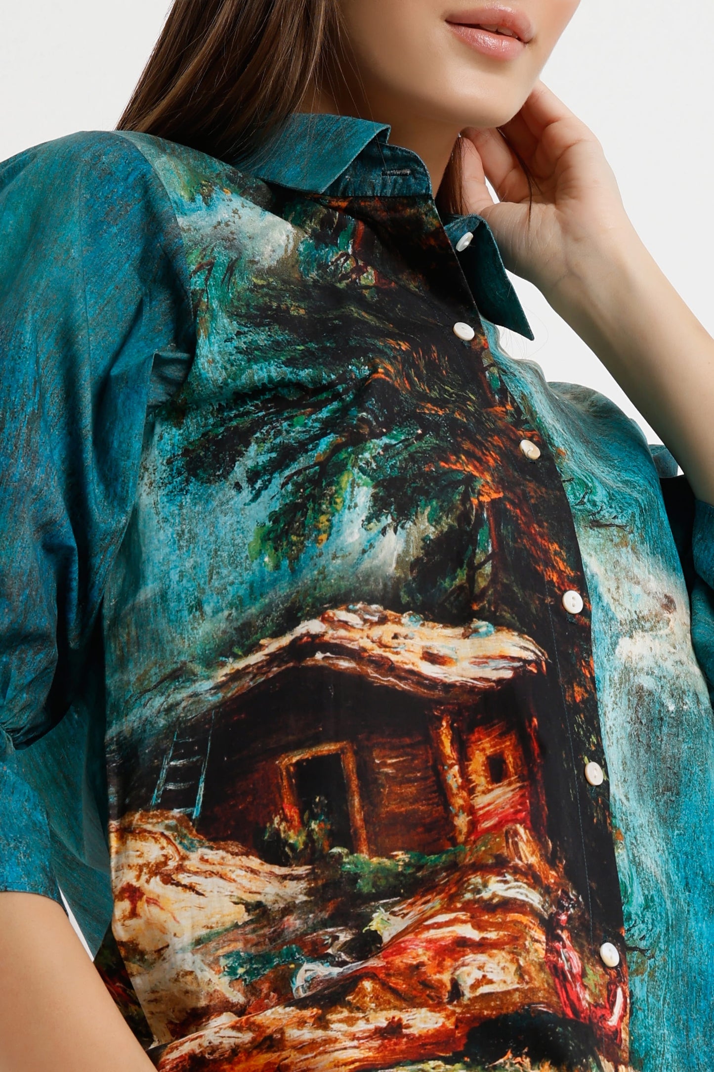 A close-up of a woman wearing a short-sleeve button-up shirt with a vibrant, painterly scenic print of a tropical cabin under palm trees in teal, brown, and orange tones.