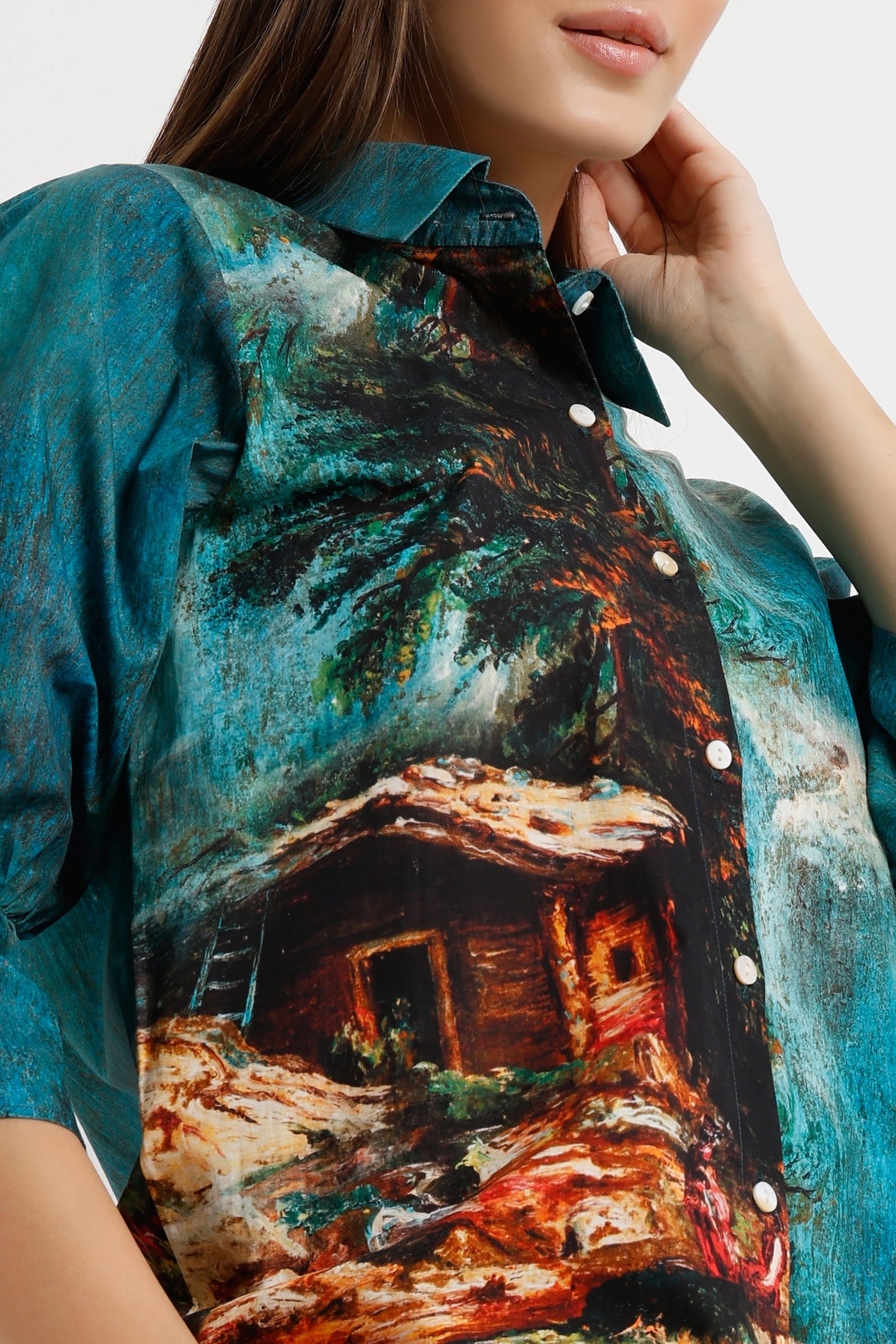 A close-up of a woman wearing a short-sleeve button-up shirt with a vibrant, painterly scenic print of a tropical cabin under palm trees in teal, brown, and orange tones.