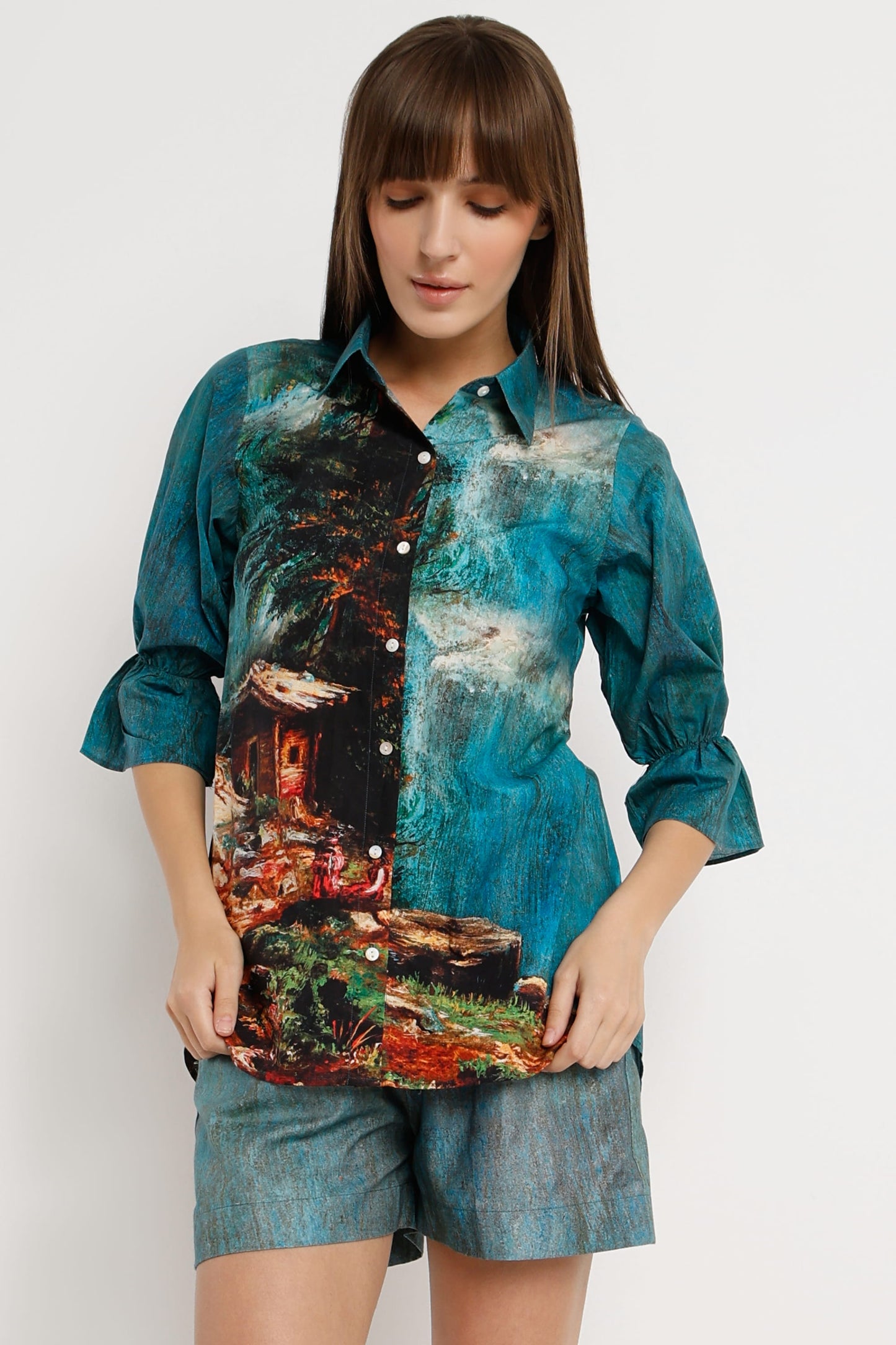A woman wearing a teal and blue printed button-up shirt with a scenic forest and waterfall design, paired with denim shorts, against a plain light gray background.