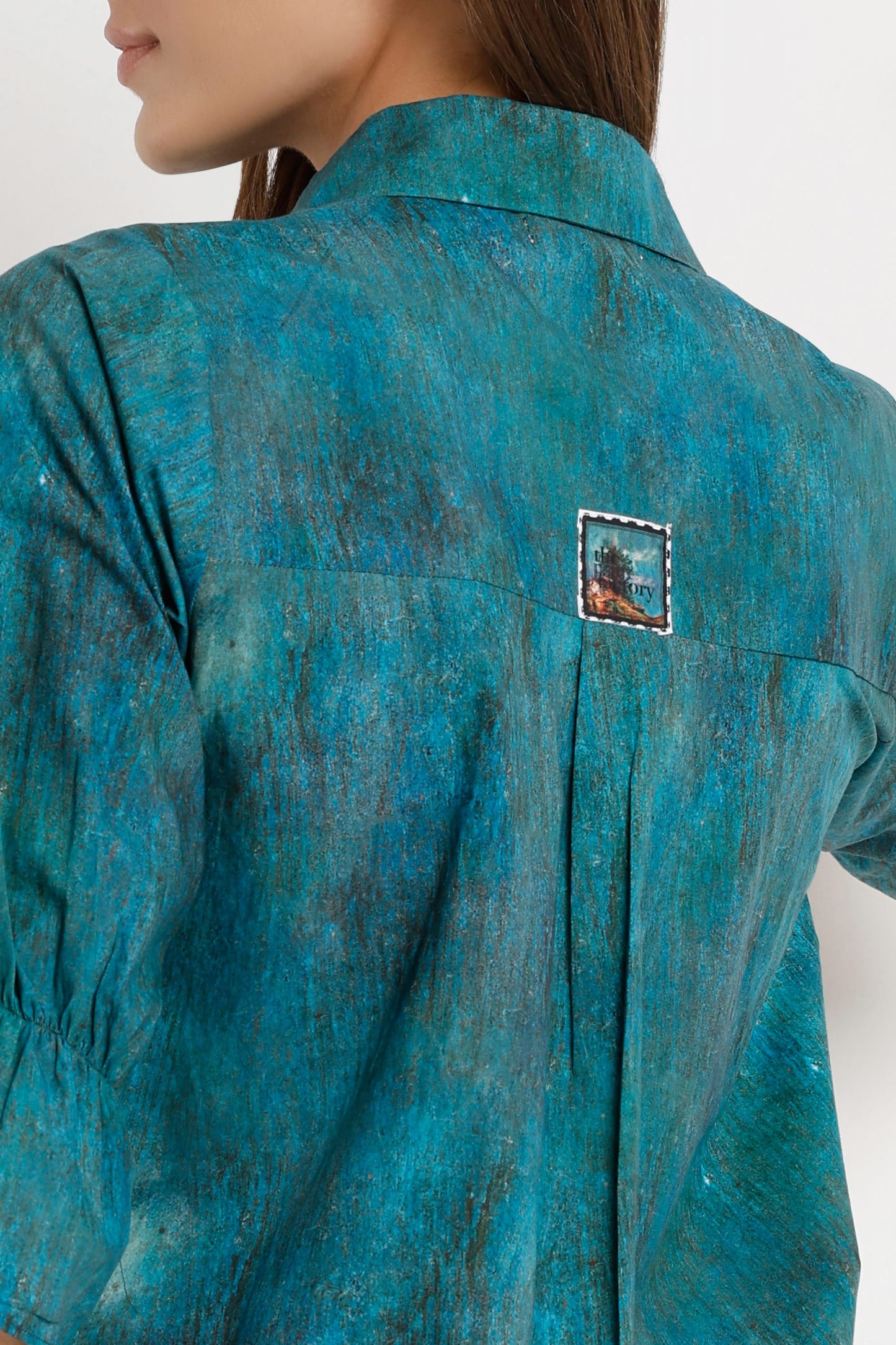 Close-up back view of a model wearing a teal blue-green textured shirt with a small square embroidered patch on the upper back.