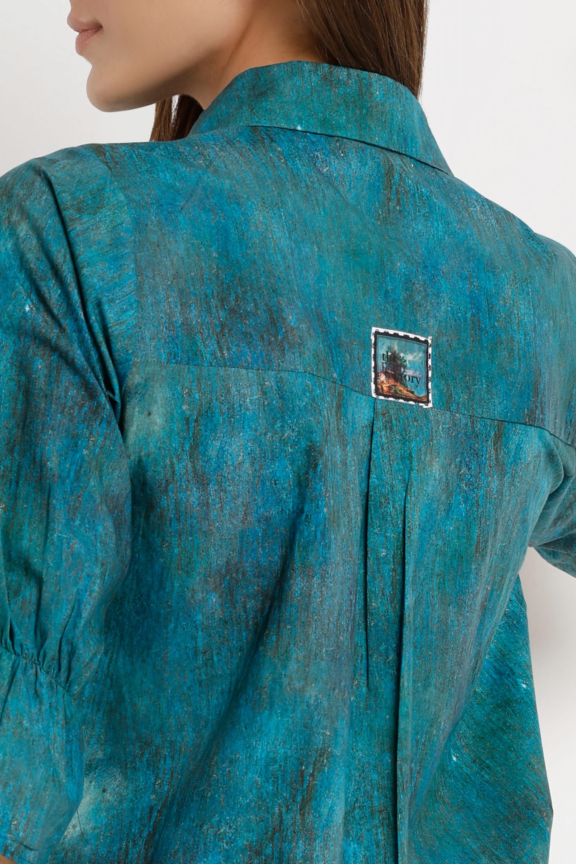 Close-up back view of a model wearing a teal blue-green textured shirt with a small square embroidered patch on the upper back.