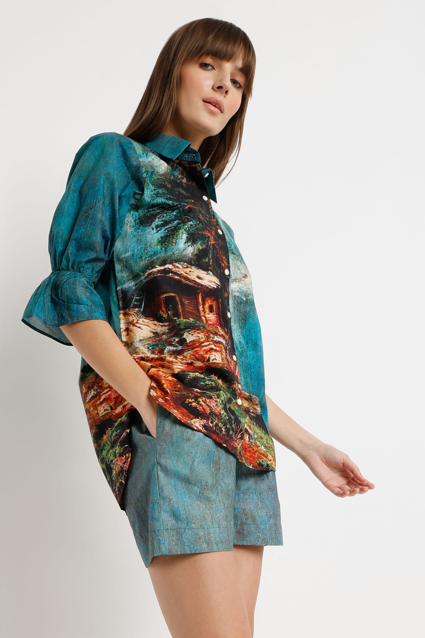 A woman modeling a short-sleeve button-up shirt with a colorful scenic print and matching shorts against a plain light gray studio background.
