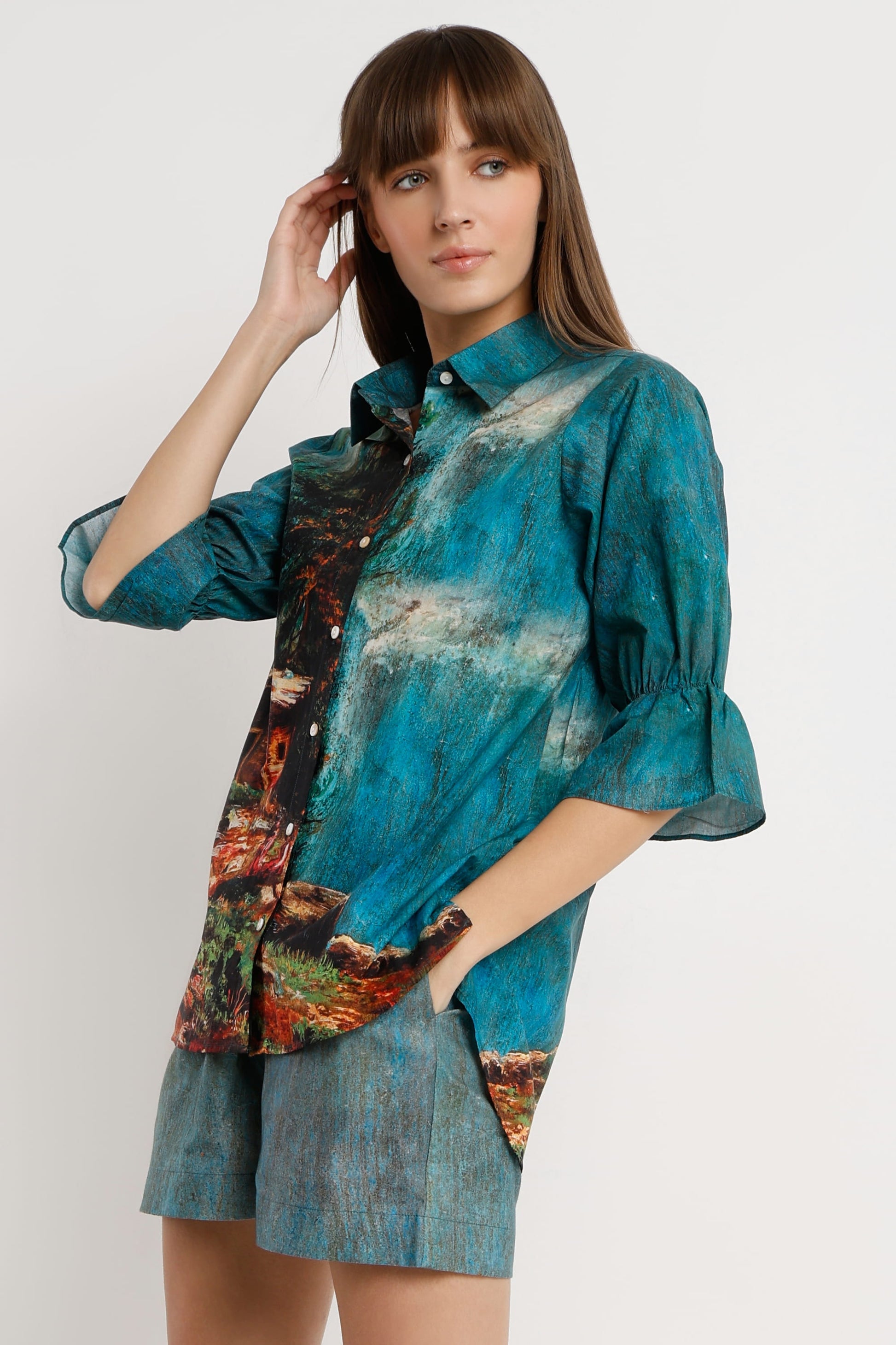 A woman modeling a teal and blue abstract print button-up shirt with ruffled sleeves, paired with matching blue shorts, against a plain light gray background.