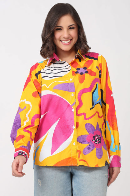 A woman wearing a long-sleeve button-up shirt with a bold, colorful retro abstract pattern in yellow, pink, orange, purple, and blue, paired with light blue jeans, against a plain white background.